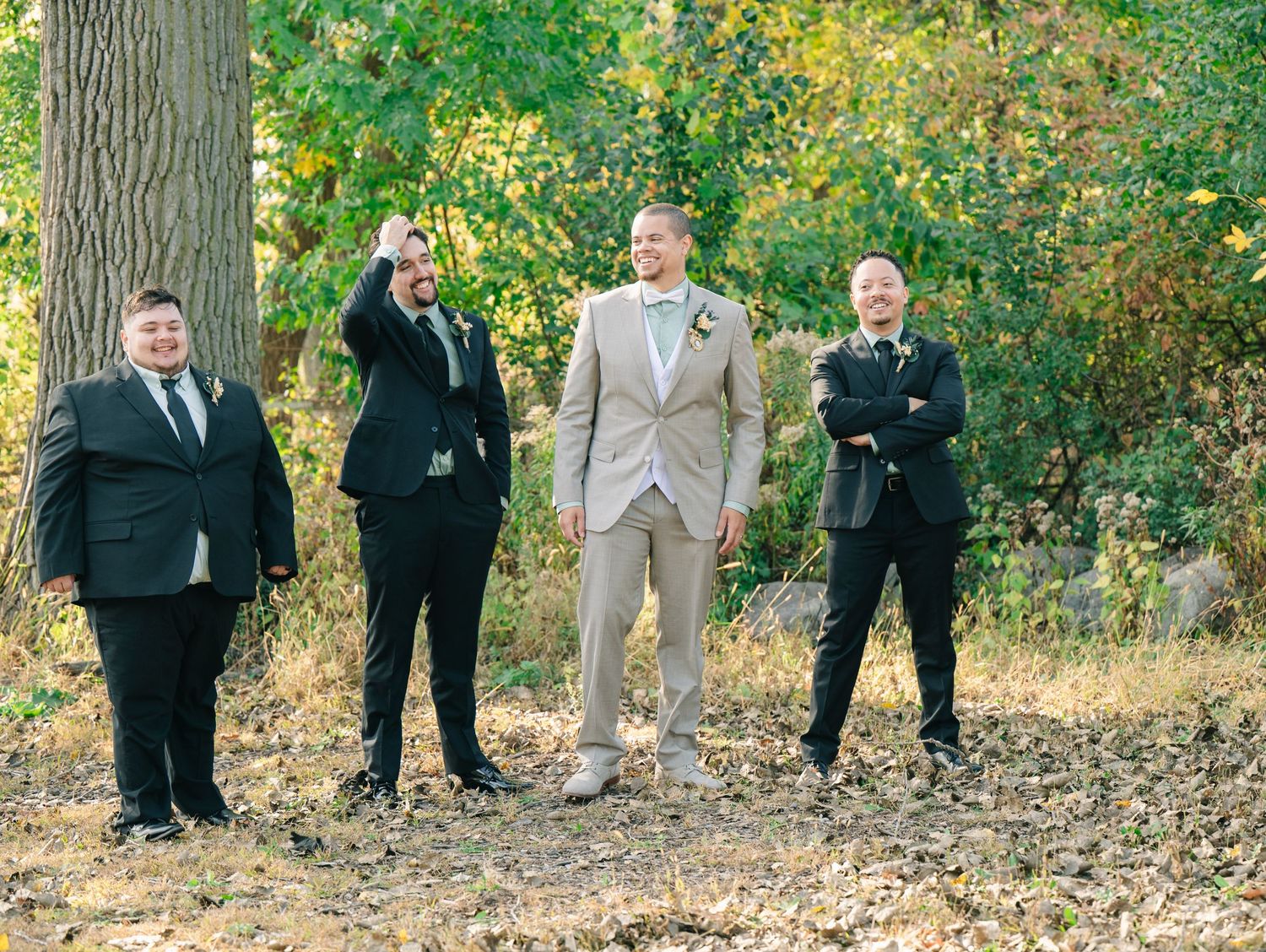 Groom in tan suit stands with groomsmen in black suits outdoors among autumn trees at a wedding.