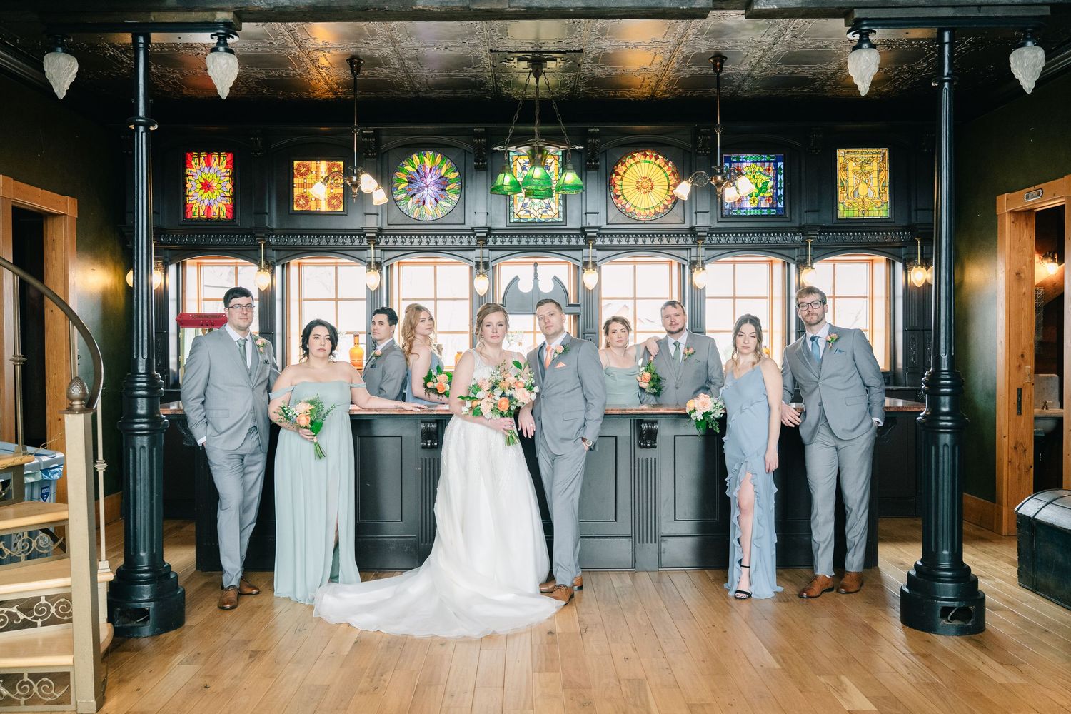 crooked willow weddings Bridal party in sage green dresses and gray suits standing in historic venue with colorful stained glass backdrop.