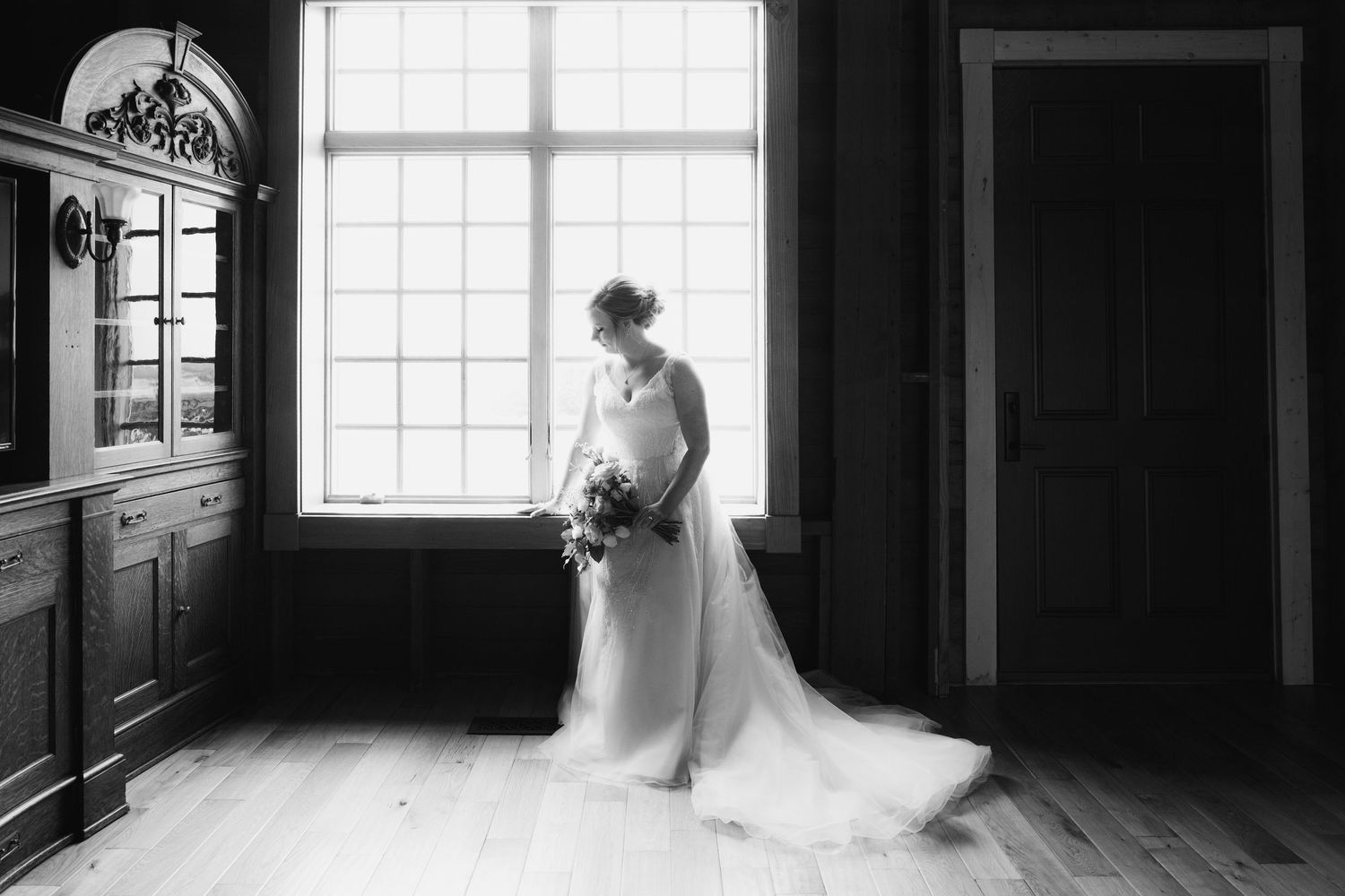 Bride in elegant white gown holding bouquet stands by large window in black and white wedding portrait. Crooked Willow Osakis