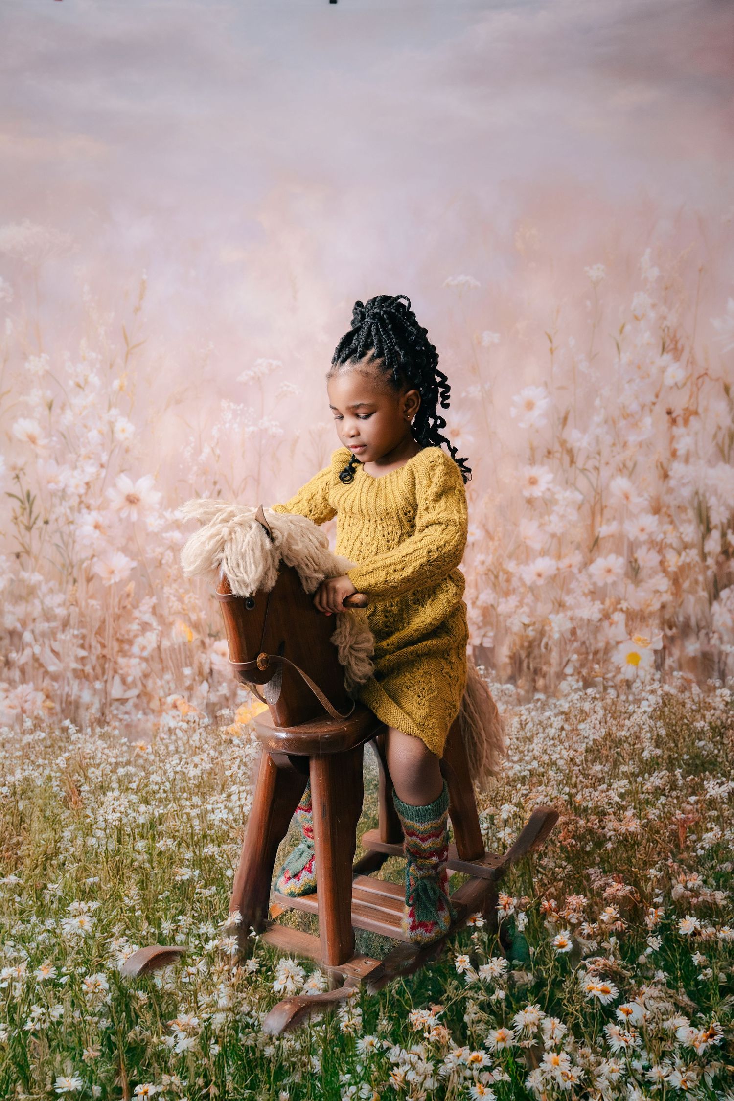 Young Black girl in yellow knit dress sits on wooden rocking horse amid dreamy floral backdrop.. Minnesota milestone photographer