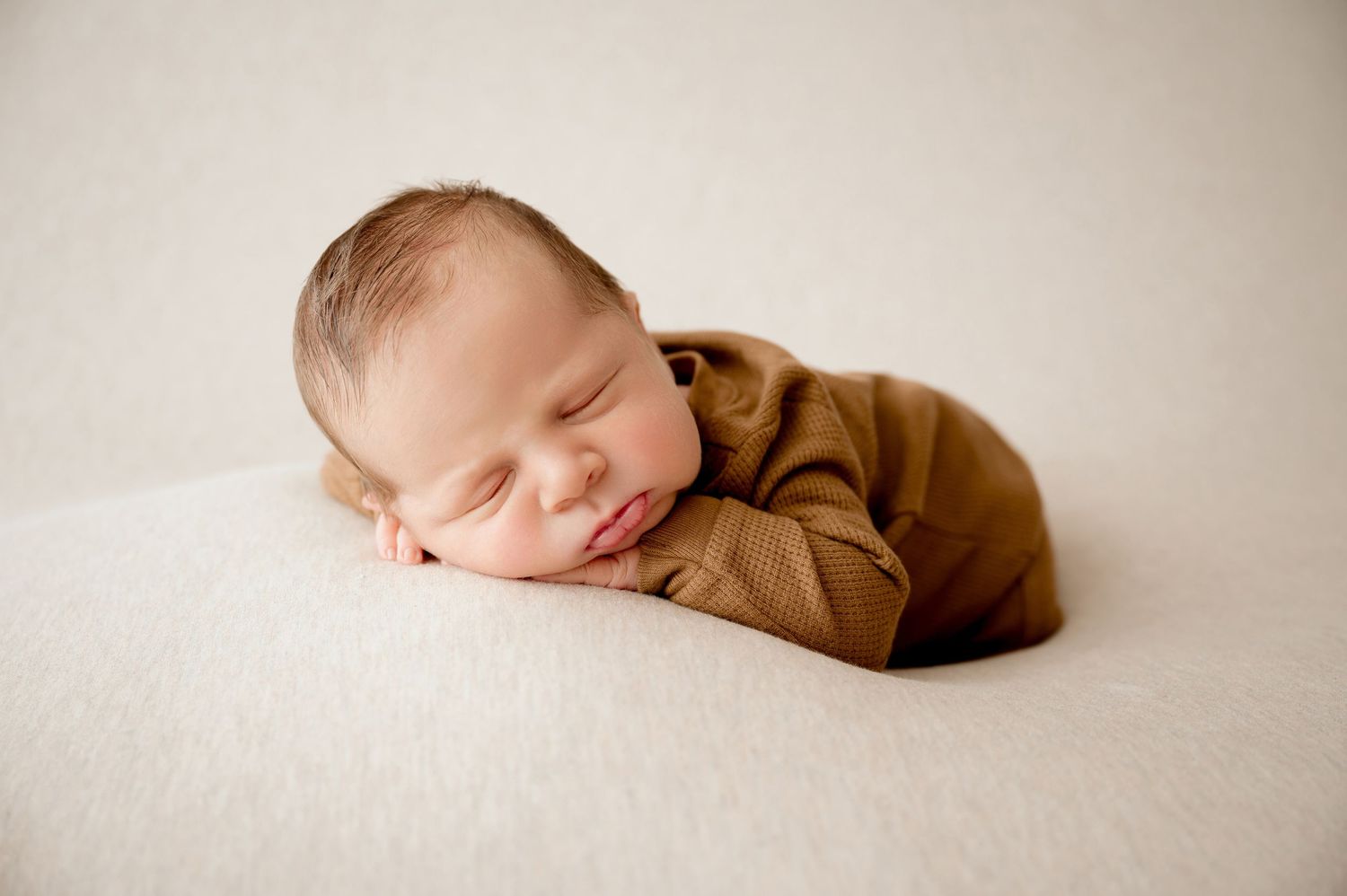 Peaceful newborn in brown outfit sleeping on cream blanket during portrait session.