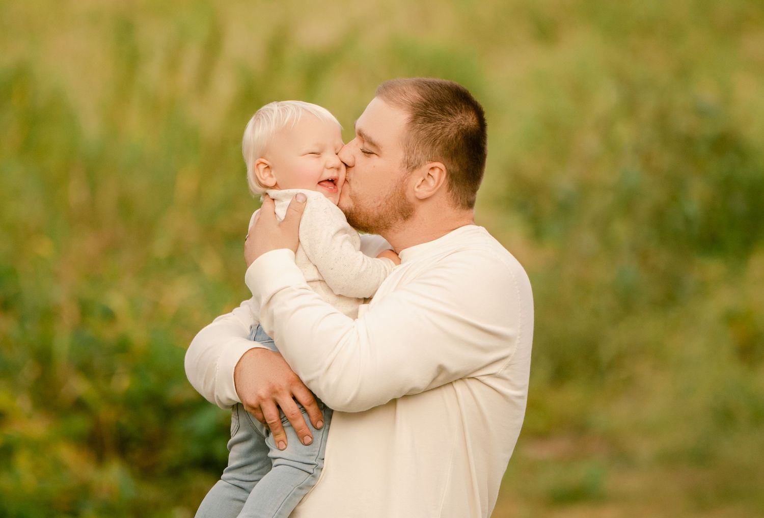 An adult in a white sweater holds and kisses a baby in light blue clothing against a blurred outdoor background.