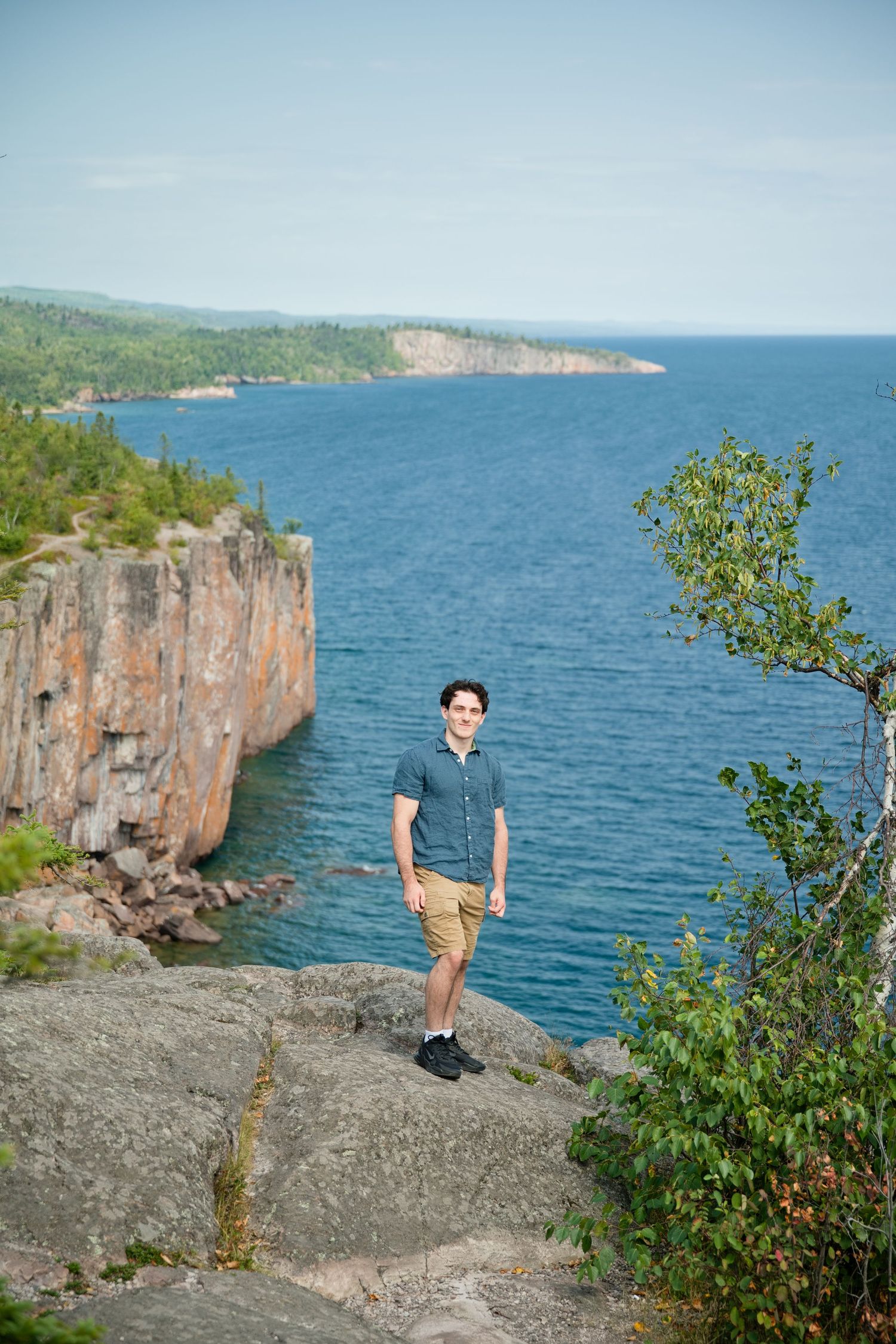 Young man in khaki shorts stands on rocky cliff overlooking stunning blue lake with forested shoreline on sunny day.
