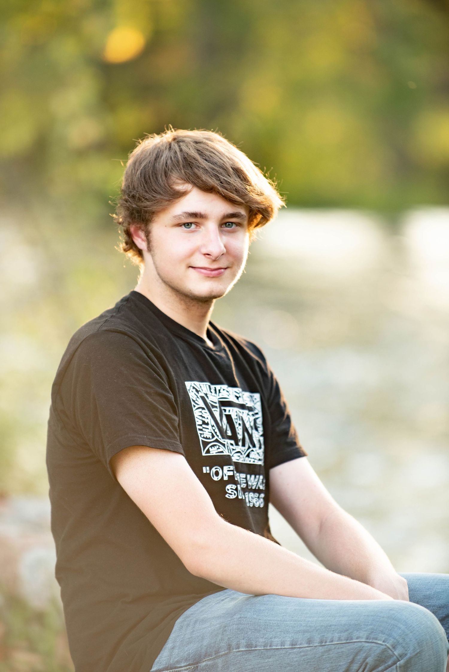 A young person in a black t-shirt sits outdoors with a soft-focus natural background.