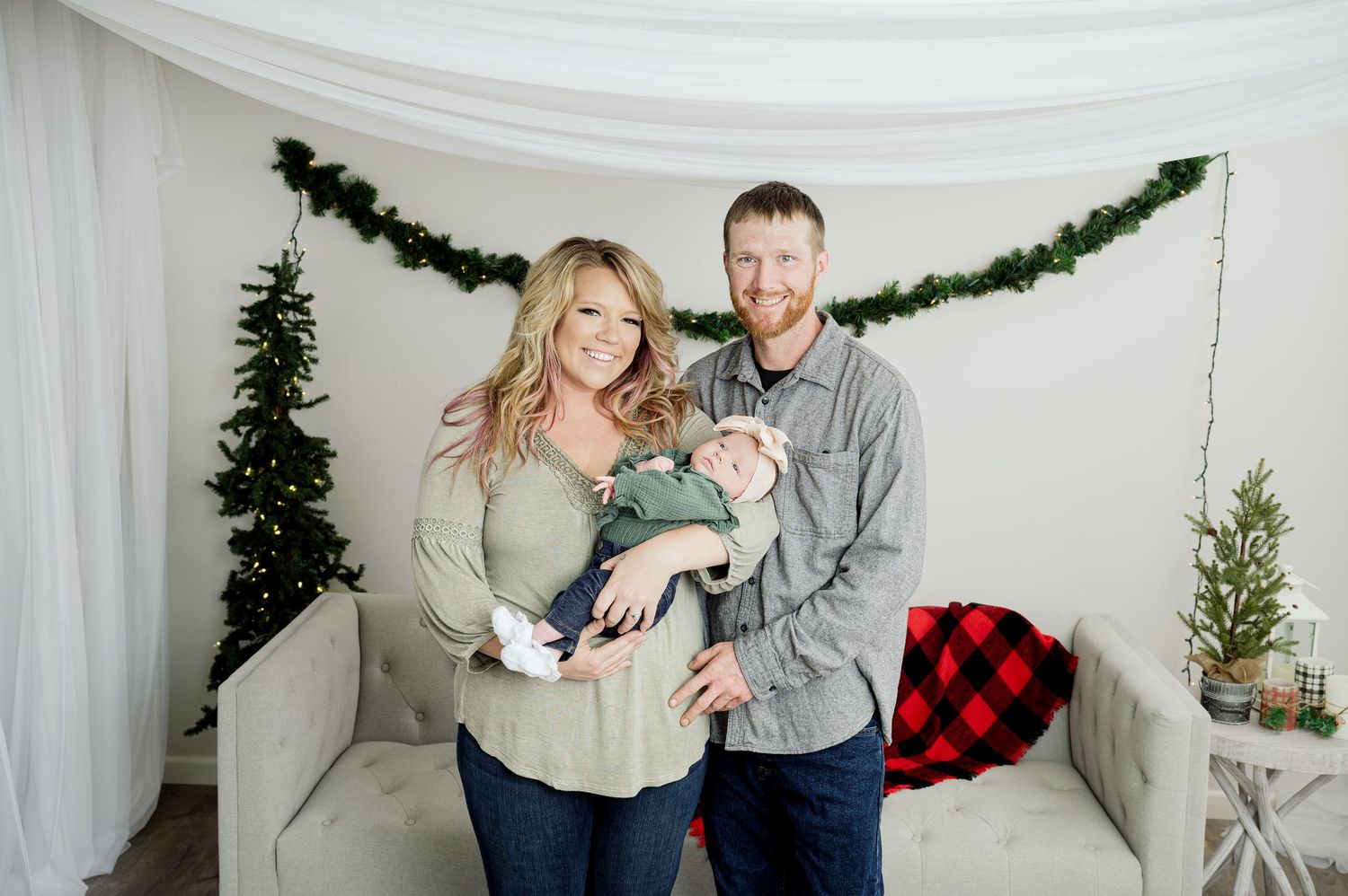 Holiday portrait with couple and small dog in cozy winter setting with buffalo plaid pillow and evergreen decorations.
