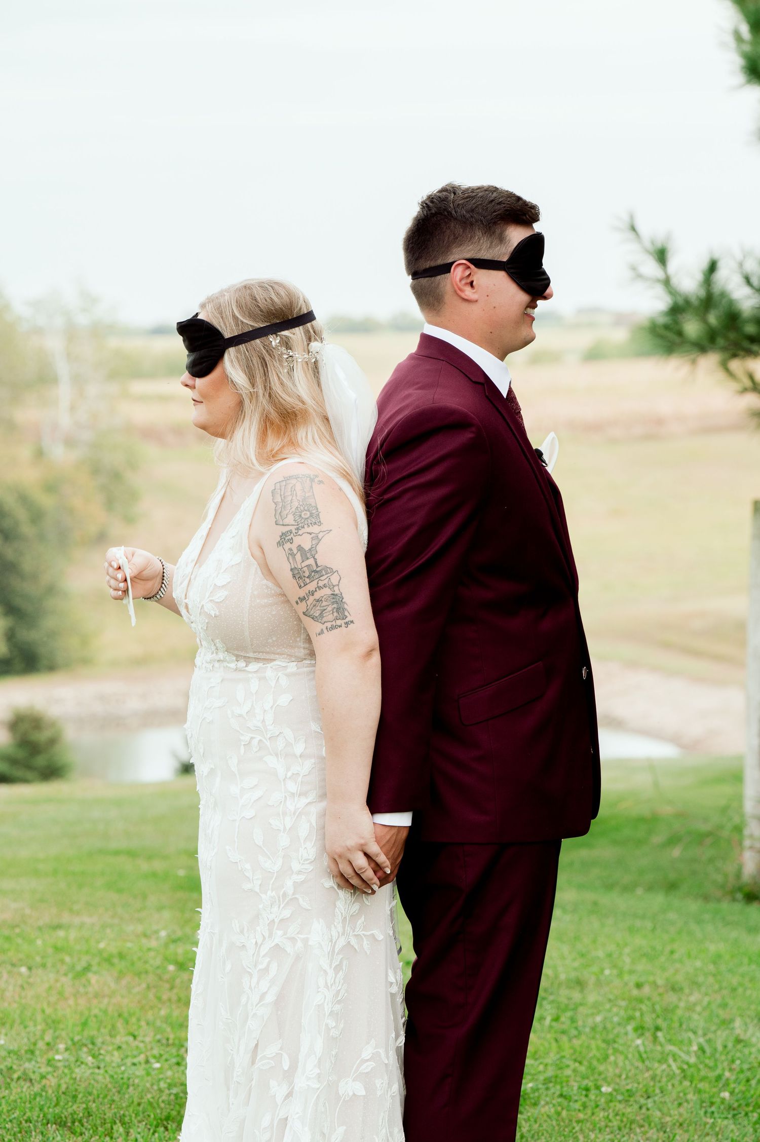 A couple in wedding attire stands back to back wearing blindfolds on a grassy outdoor setting.
