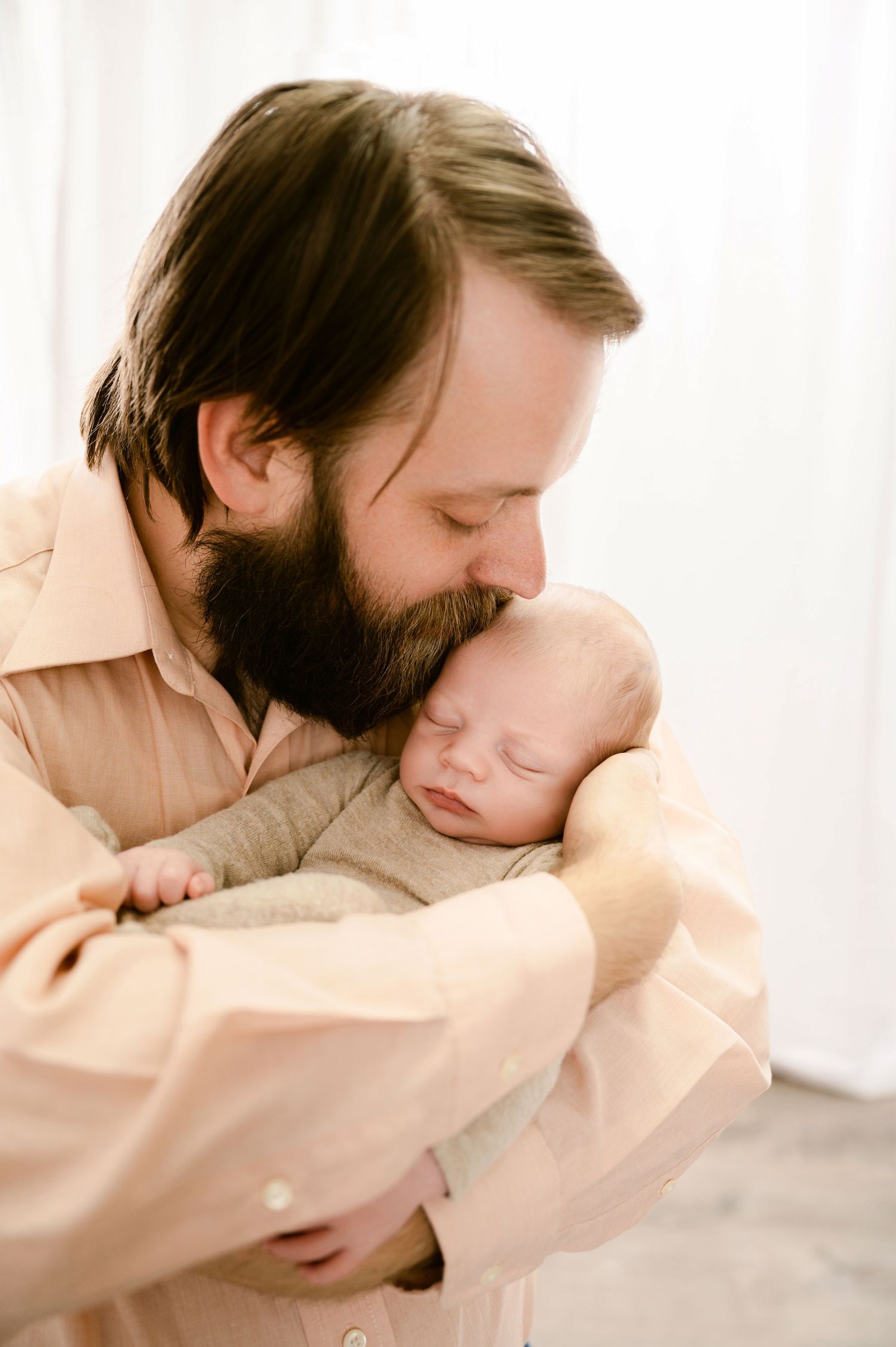 A parent wearing a beige shirt tenderly kisses their sleeping newborn baby while holding them in their arms.