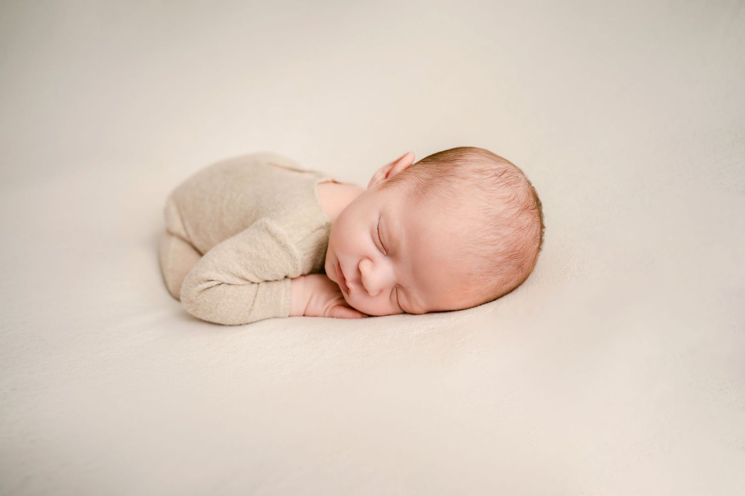 Peaceful sleeping newborn baby wearing cream colored onesie rests curled up on soft beige backdrop.
