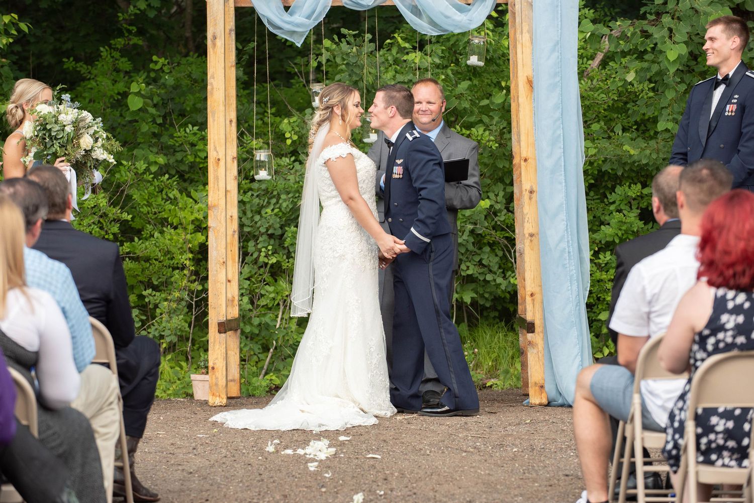 An outdoor wedding ceremony takes place under a wooden arch decorated with flowing white fabric in a natural setting. Zimmerman MN Weddings