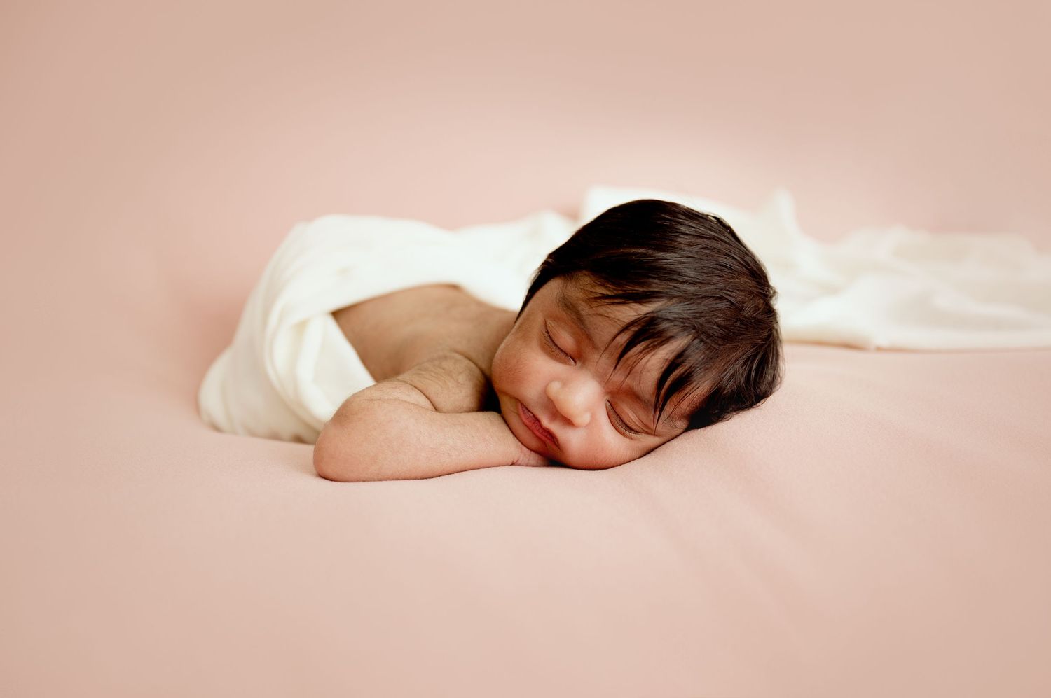 Peaceful newborn baby sleeping on a pink blanket, partially wrapped in a soft white cloth.