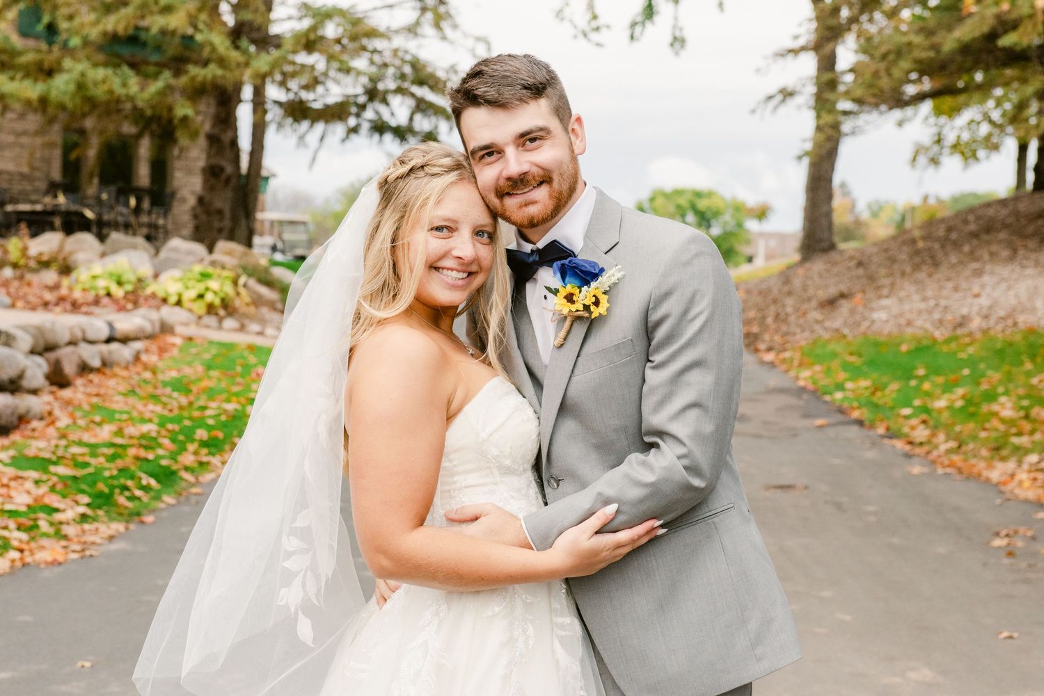 Central MN Wedding Smiling bride in white gown and veil with groom in gray suit and floral boutonniere posing outdoors on their wedding day.