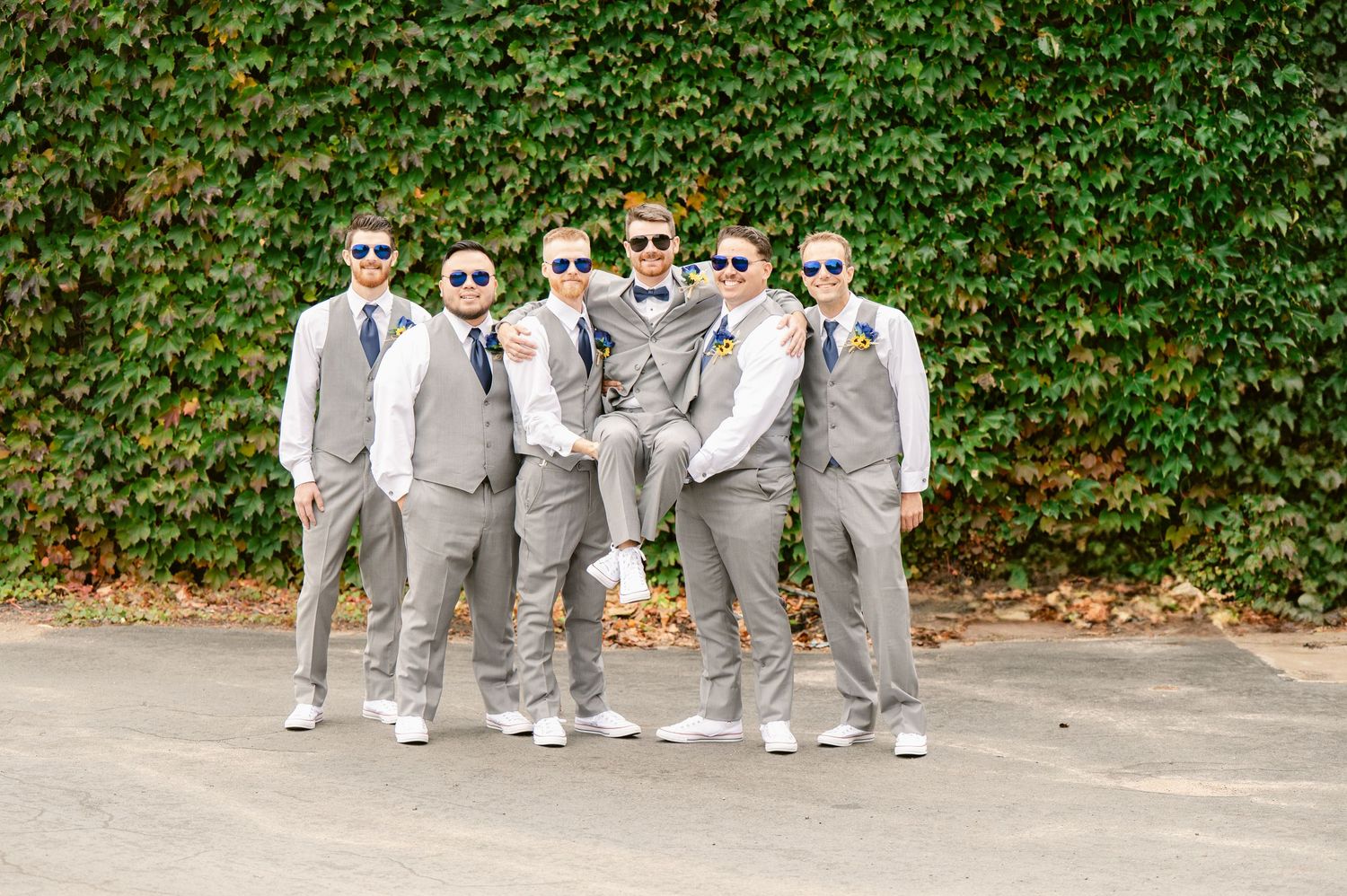 Central MN Wedding Photos | Five groomsmen in matching gray suits and sunglasses pose playfully outdoors against a lush green ivy wall.