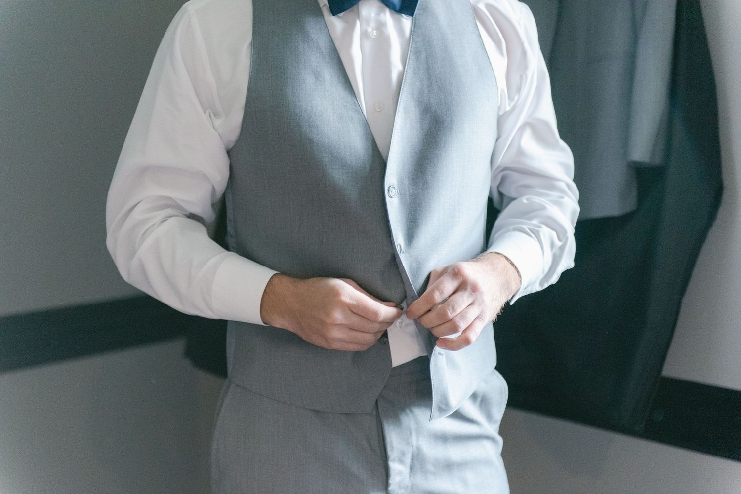 Central MN Wedding Photos | Groom buttoning his grey vest while wearing a white shirt and navy bow tie on wedding day.