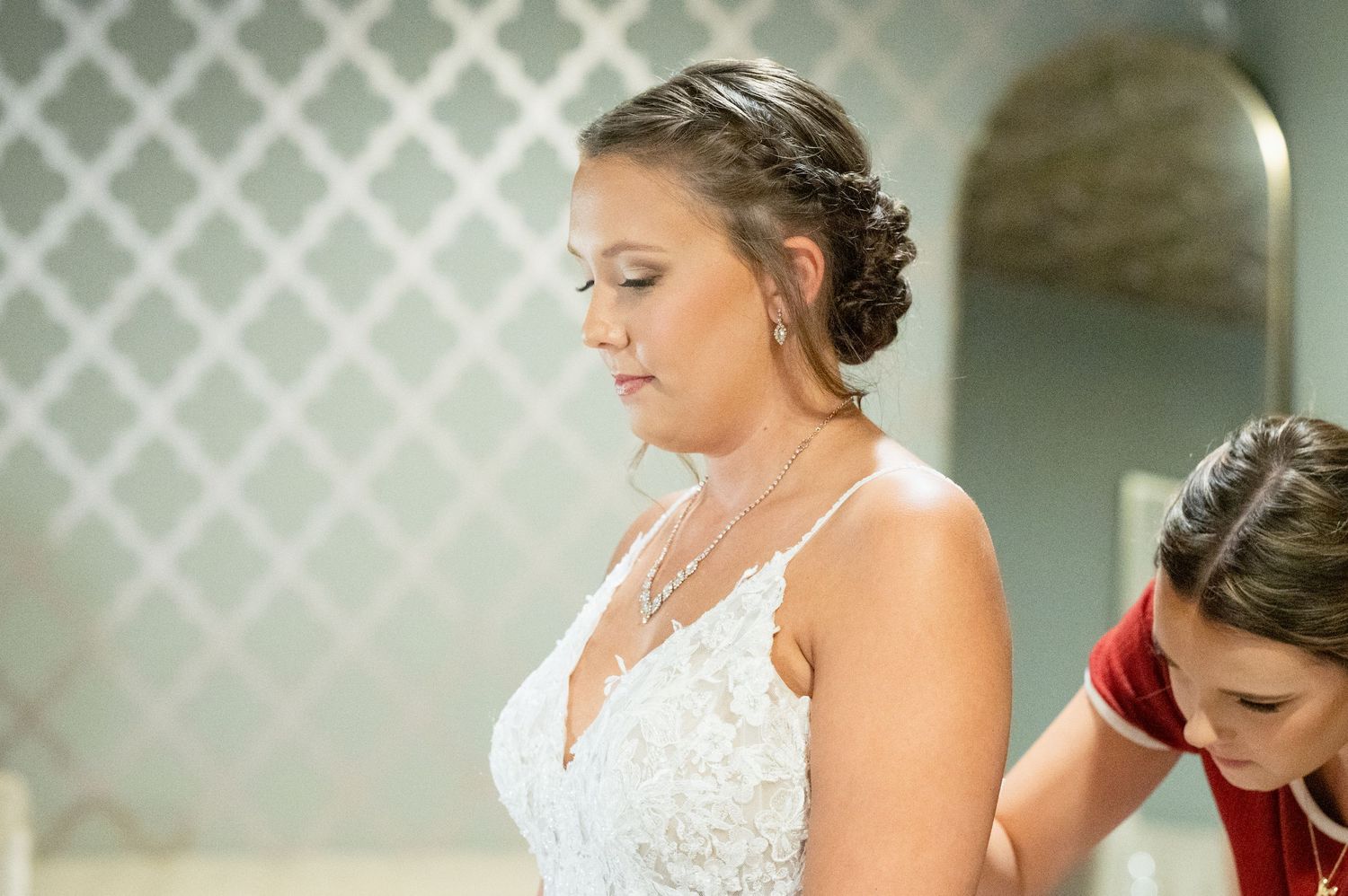Bride in lace wedding gown having dress fastened by bridesmaid in elegant bridal preparation scene.