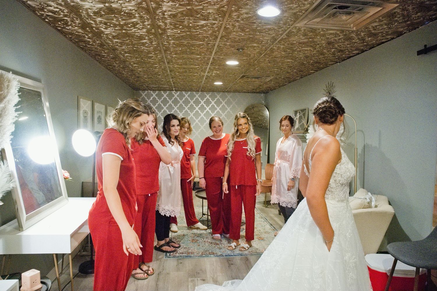 Bridesmaids in red outfits react emotionally as bride reveals wedding dress in elegant dressing room.