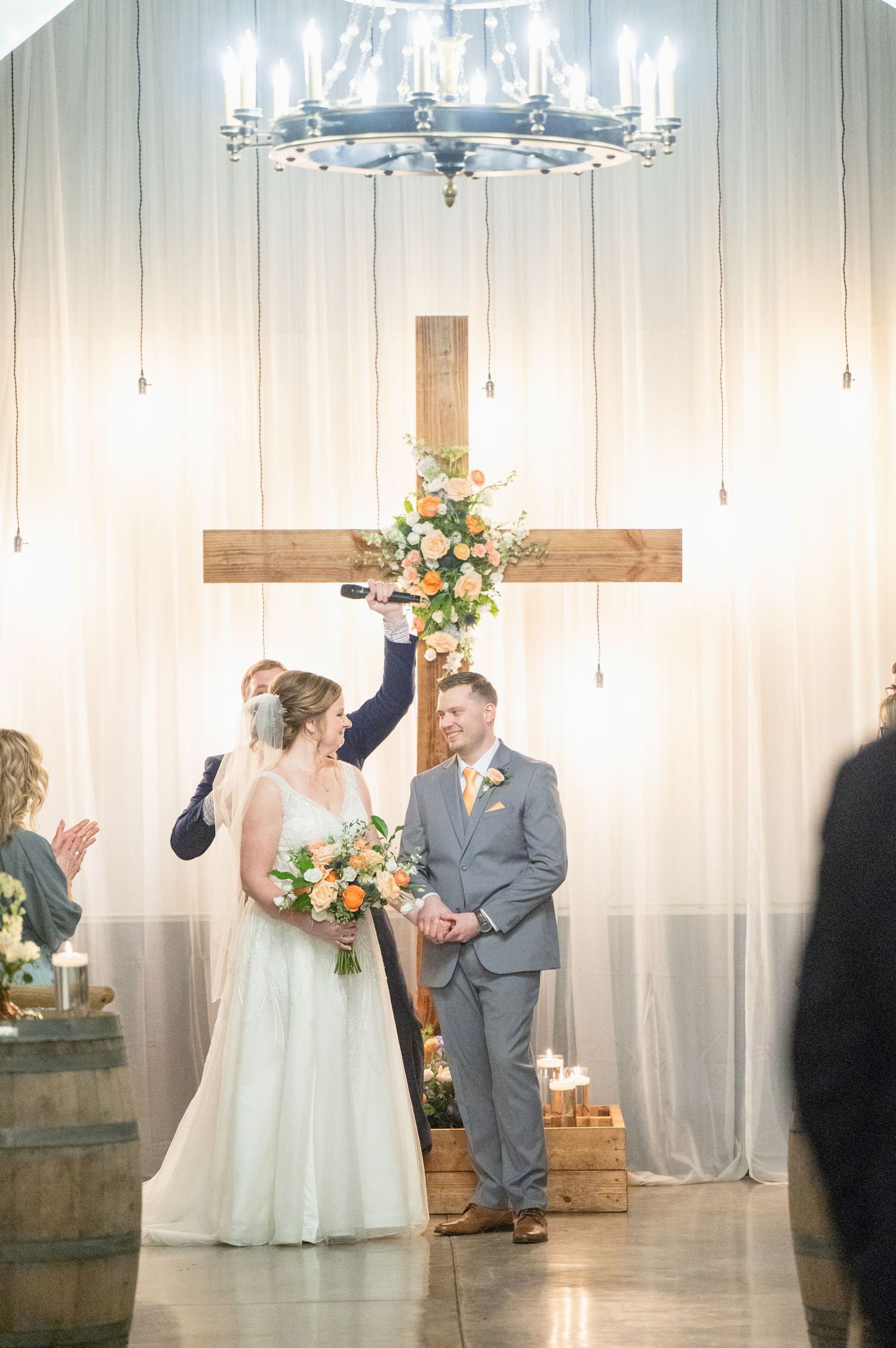 Bride and groom stand before a wooden cross adorned with flowers during an indoor wedding ceremony.