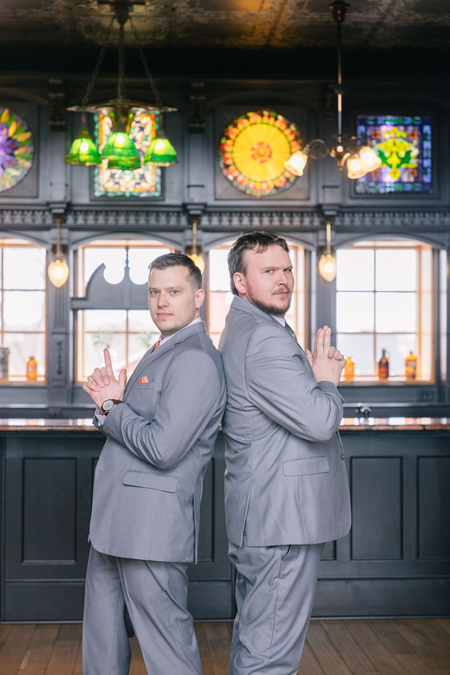 Two groomsmen in gray suits stand back-to-back with serious expressions in an elegant bar venue.