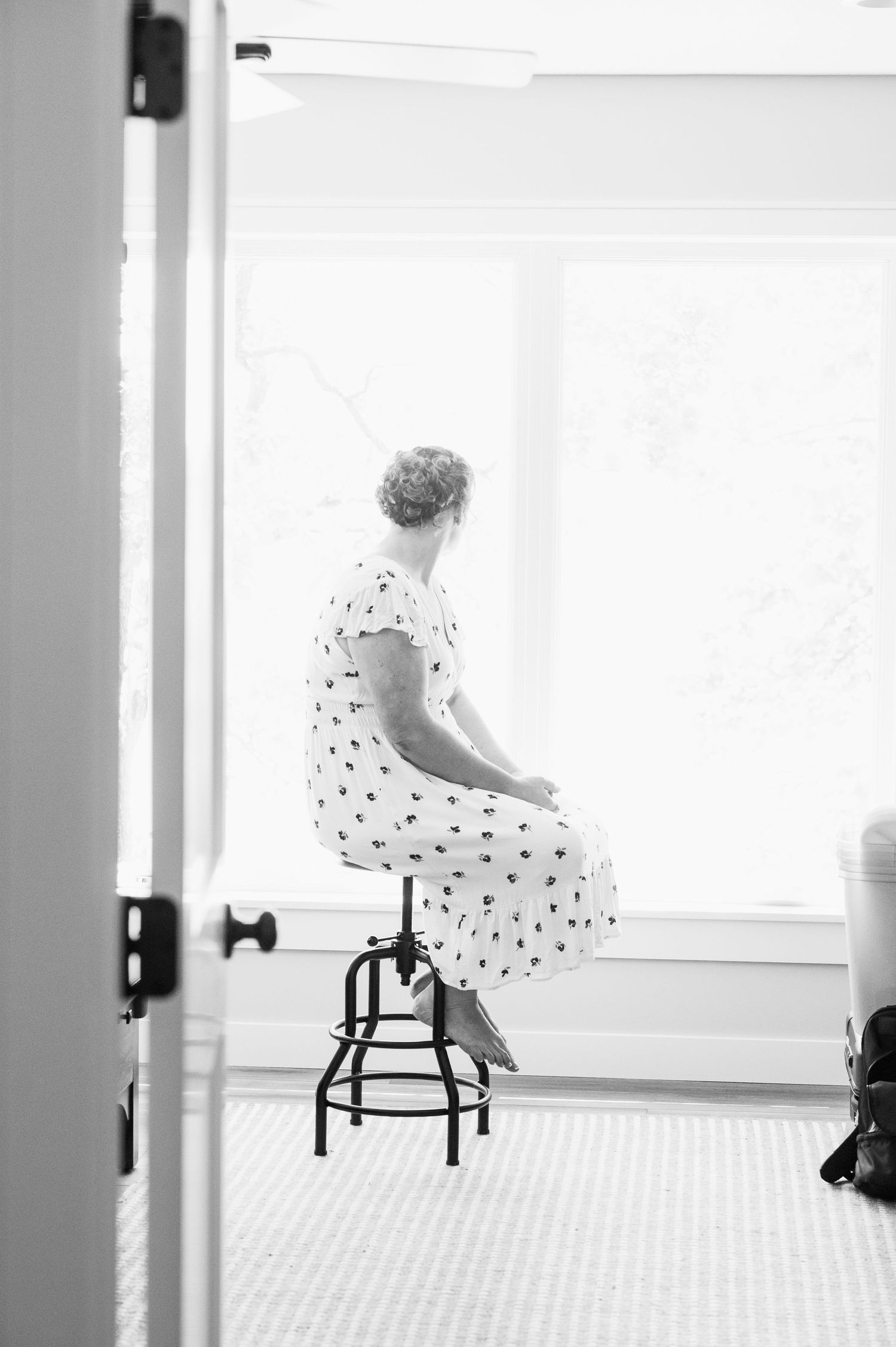 Elderly woman in floral dress sits on metal stool facing large window in black and white photo.