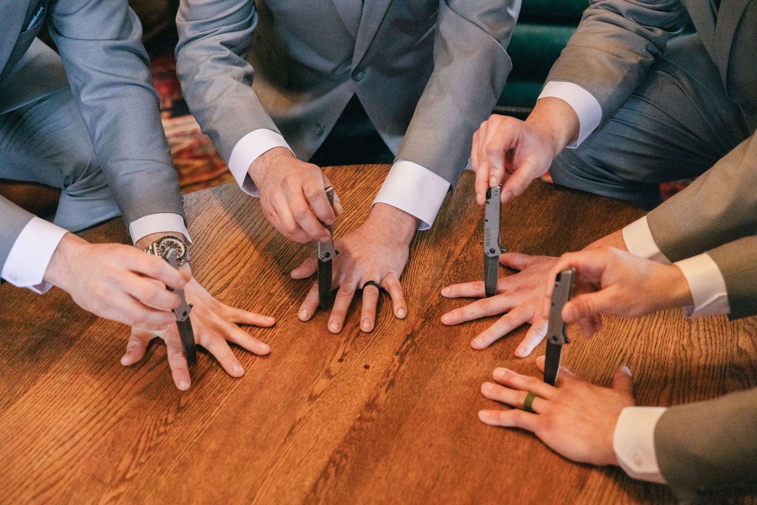Groomsmen in gray suits place hands on a wooden table holding cufflinks during wedding preparations.