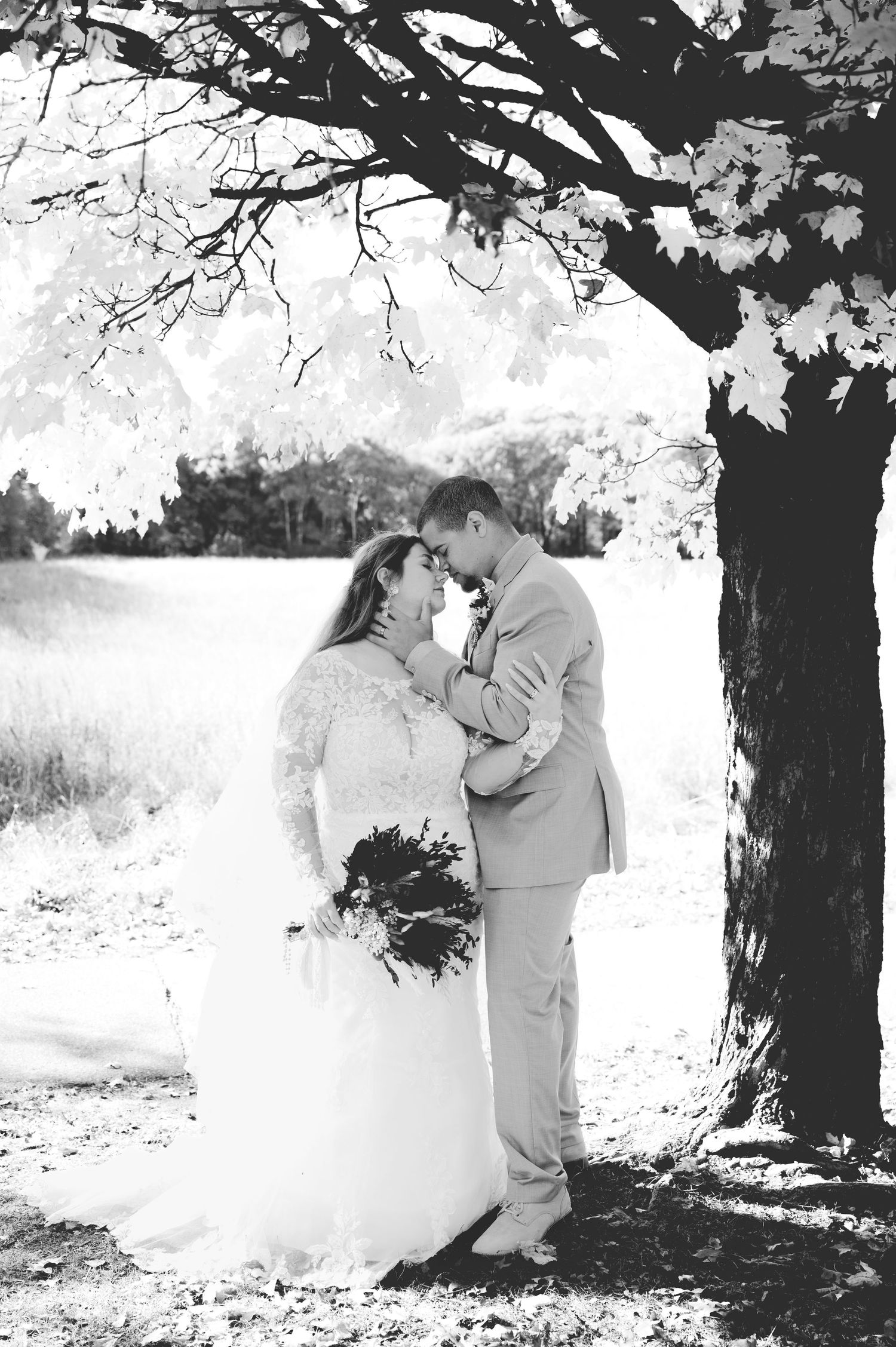 Black and white wedding photo of bride and groom embracing under a tree, bride holding bouquet.