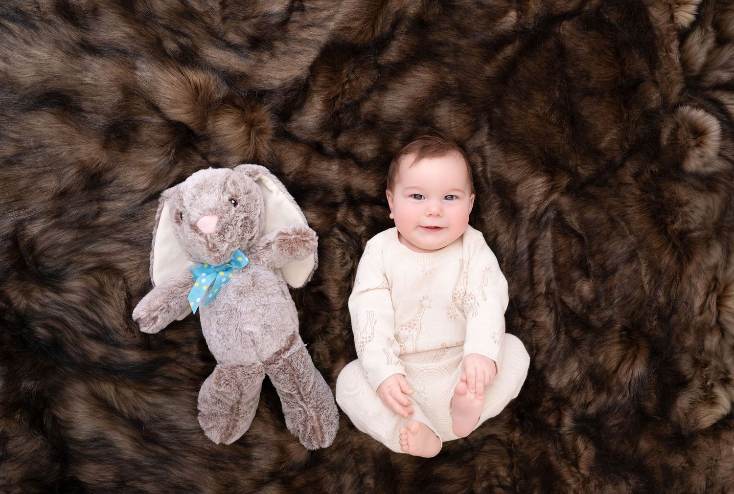 Baby in white outfit lying next to stuffed bunny toy with blue bow on brown fur blanket during professional photo session.. Minnesota milest