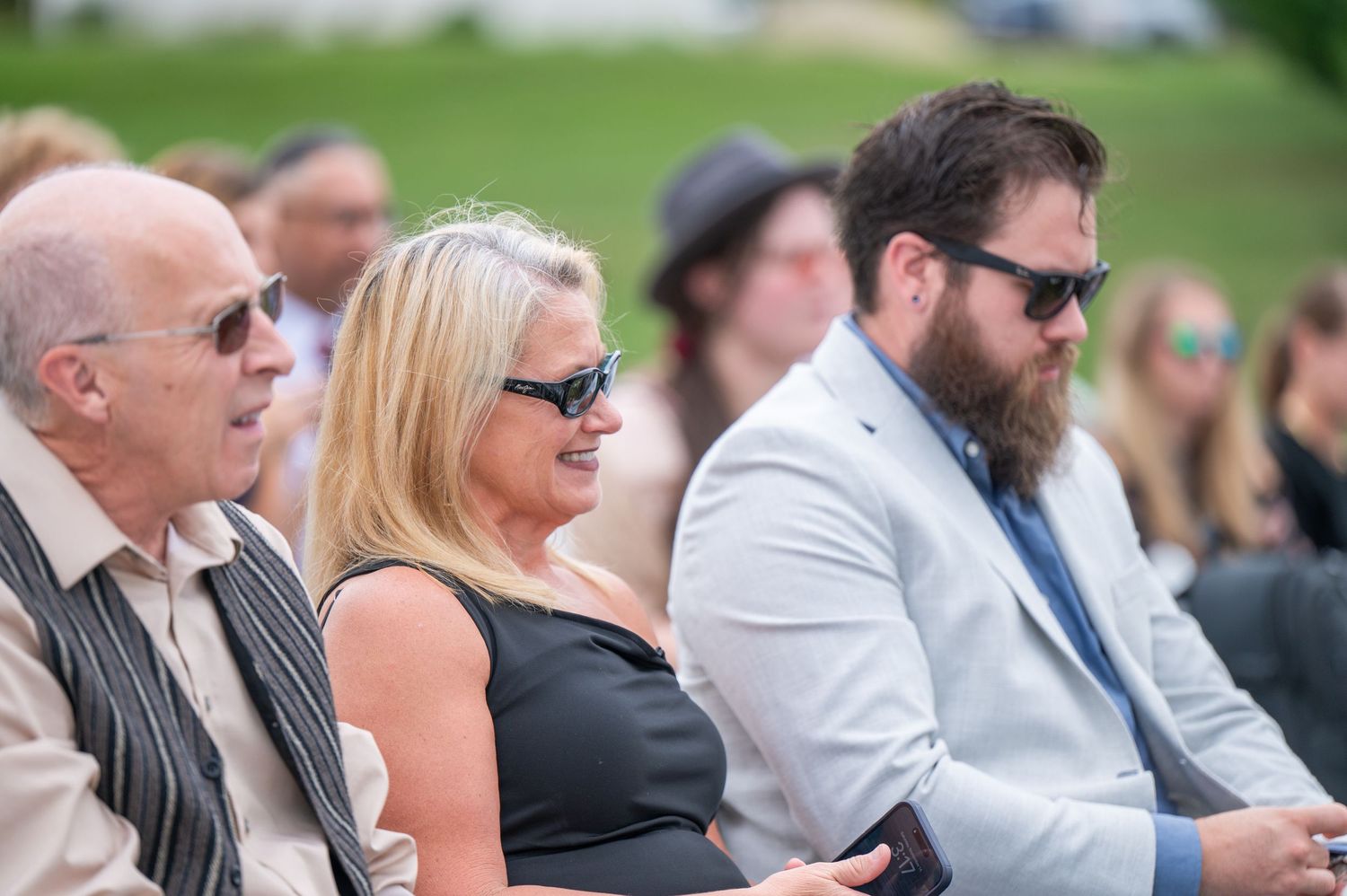 Wedding guests in sunglasses sit attentively during an outdoor ceremony.