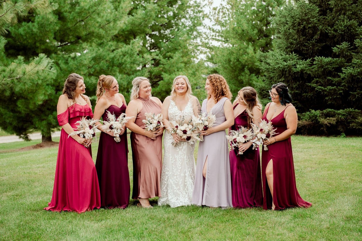 Bridal party poses together outdoors wearing mix of burgundy, blush, and neutral colored gowns.