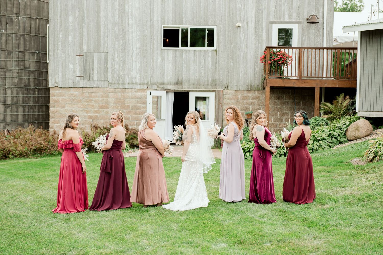 Wedding party poses in front of rustic barn with bridesmaids wearing shades of burgundy and blush gowns.