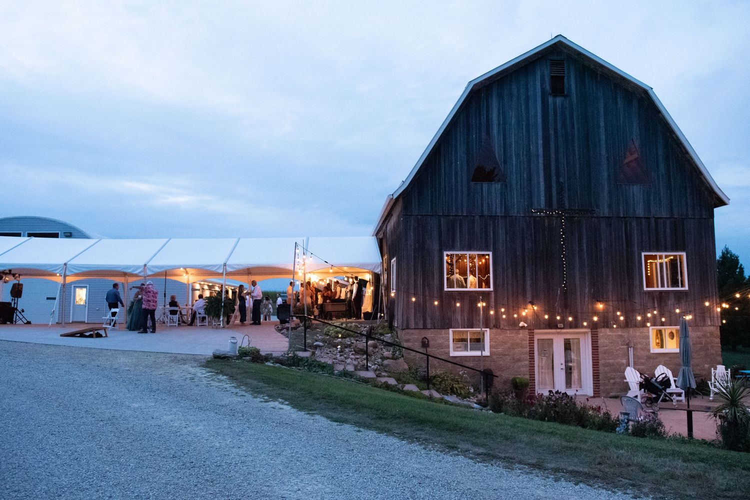 Rustic barn wedding venue at dusk with white tent reception area and warm glowing lights along stone pathway.