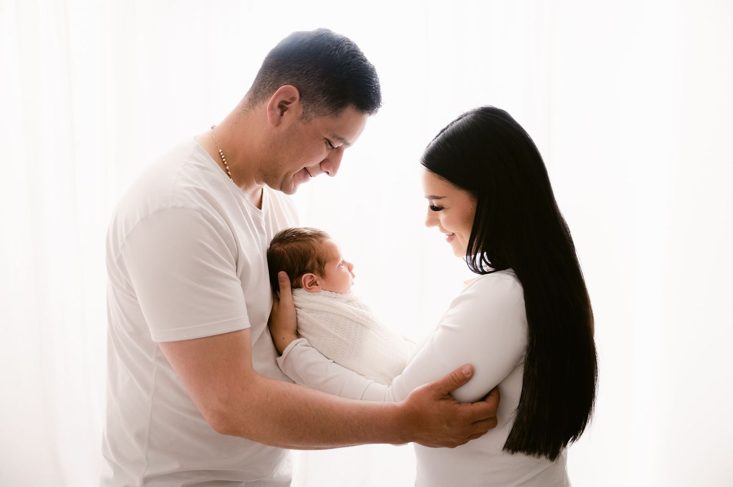 New parents in white outfits lovingly cradling their newborn baby by bright window.
