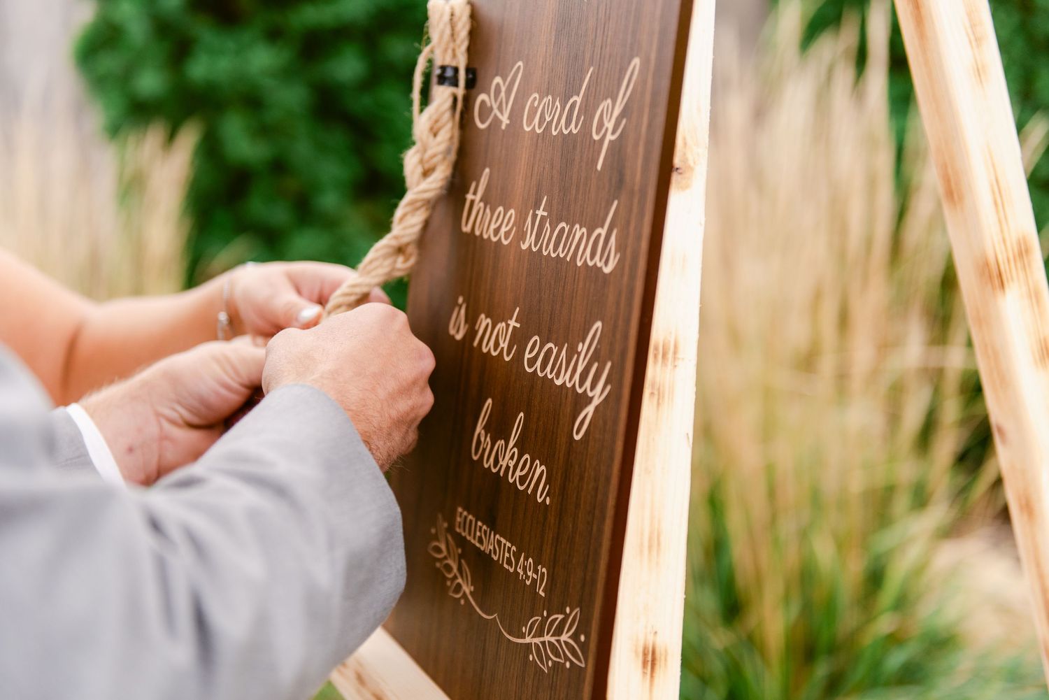 Central MN Wedding Photos - Hands braiding rope on a wooden wedding sign reading a cord of three strands is not easily broken.