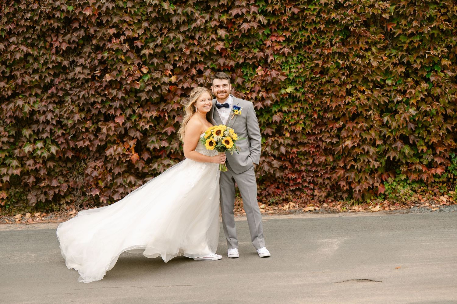 Central MN Wedding Photos | Smiling bride and groom pose before vibrant red ivy wall, bride holding bright sunflower bouquet.
