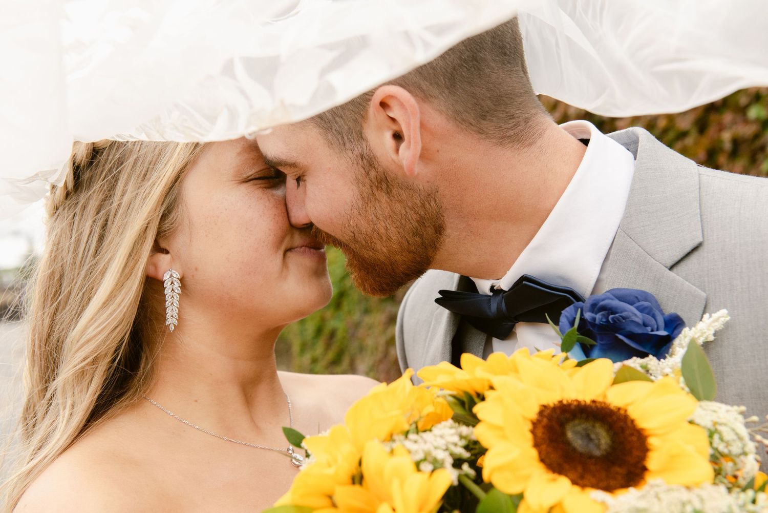 Central MN Wedding Photos | Bride and groom share a romantic kiss under a veil with sunflower bouquet in foreground.