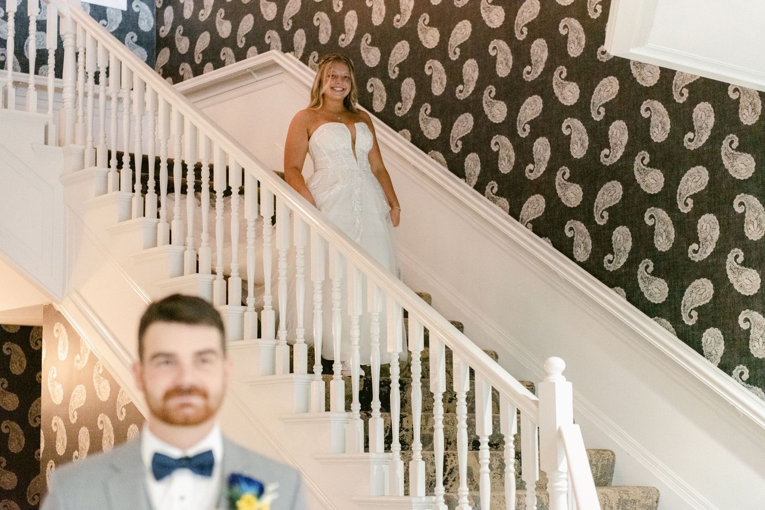Central MN Wedding Photos | Bride in white gown descending elegant staircase with paisley wallpaper as groom waits below.