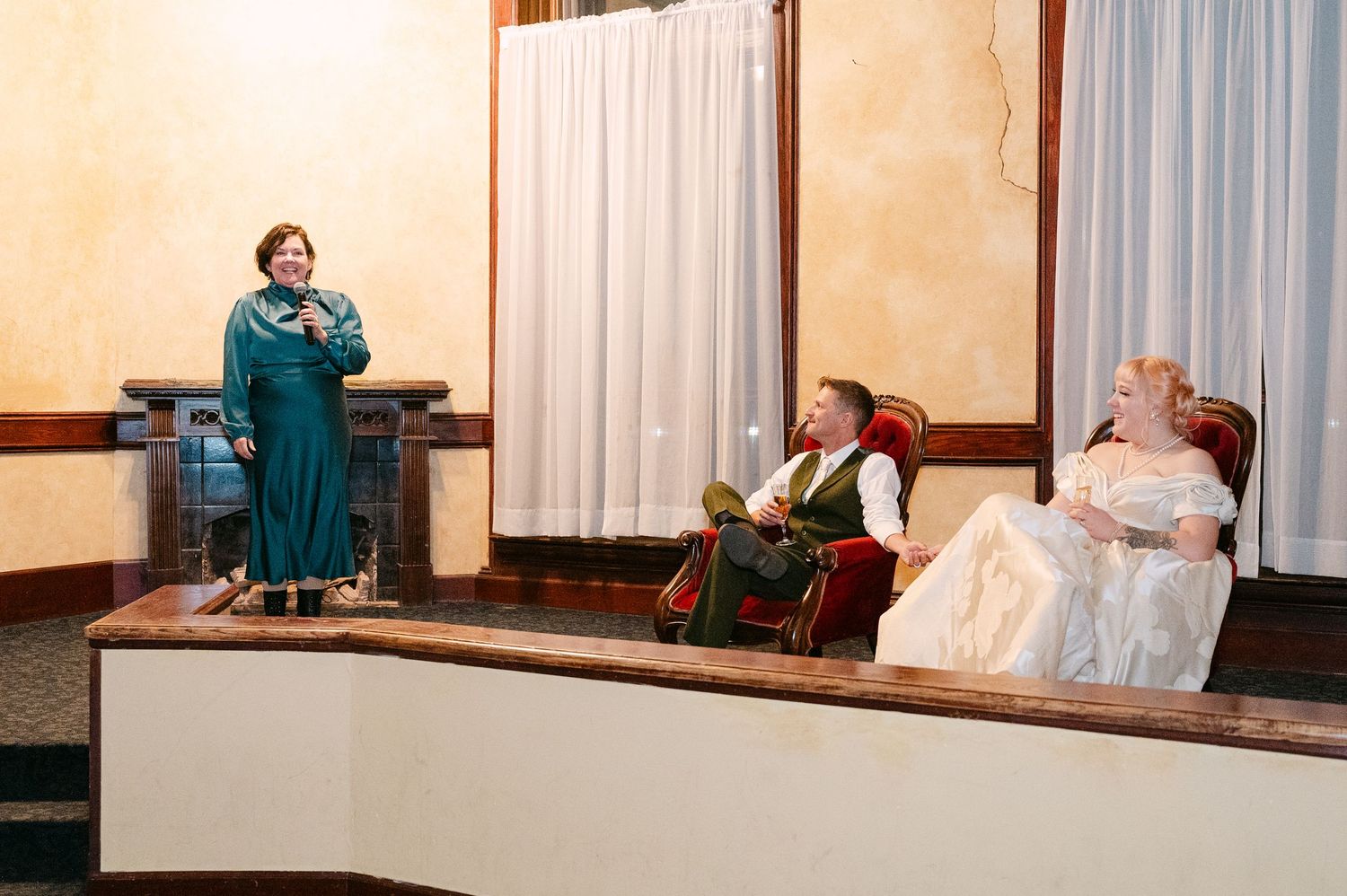 Central MN Wedding Photos | Wedding reception speech by woman in teal dress while bride and groom sit on ornate chairs.