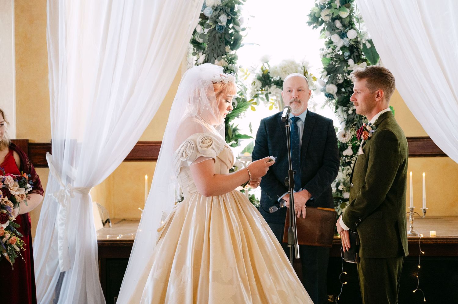 Central MN Wedding Photos | Bride in golden ballgown reading vows to groom at decorated indoor wedding ceremony altar.