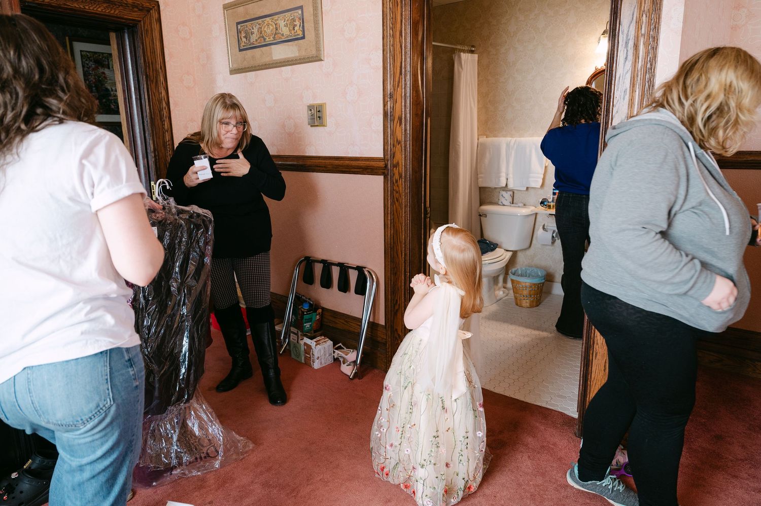 Central MN Wedding Photos | Women and a young girl in a white dress gathered in a pink hallway near a bathroom.