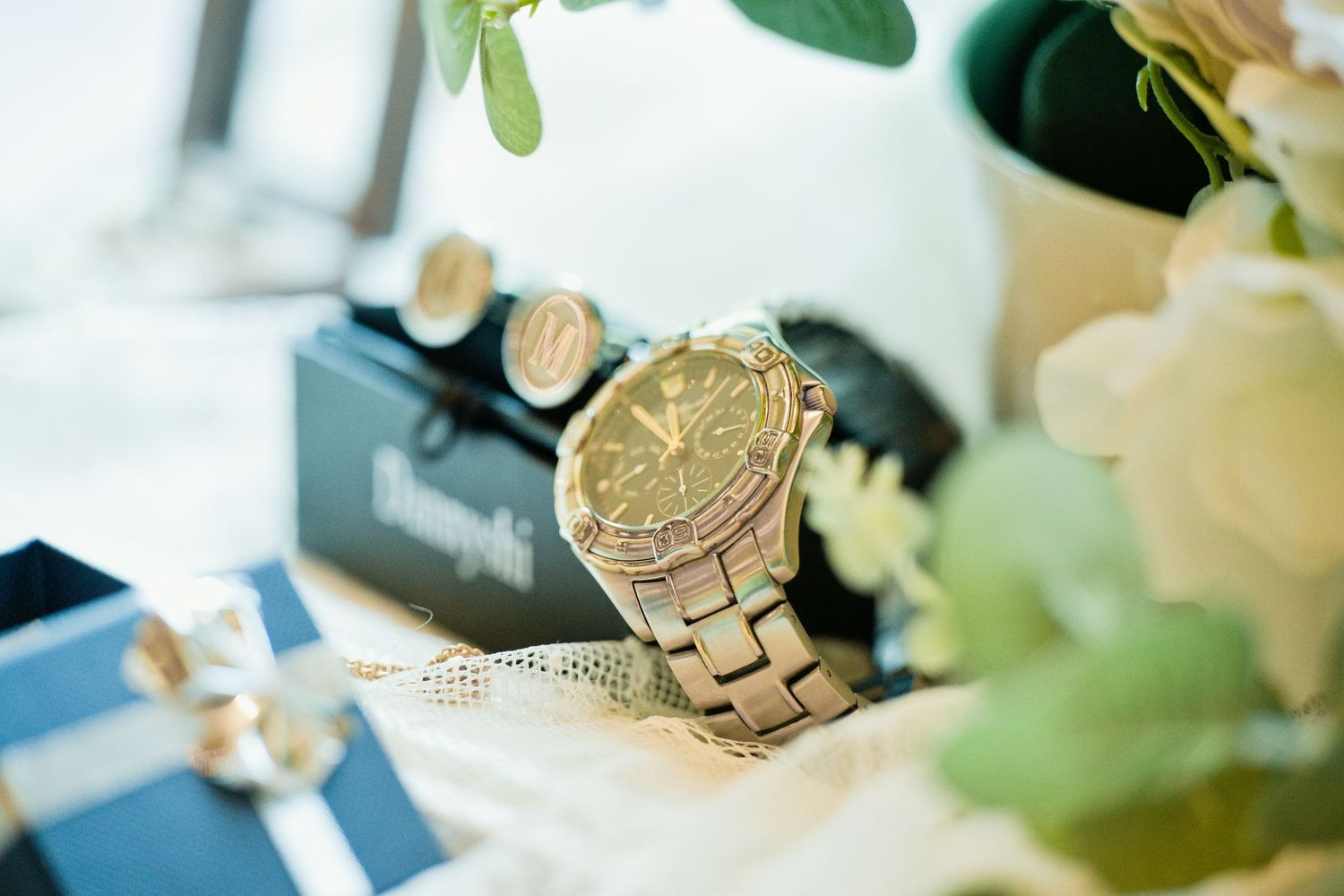 Gold luxury watch with matching cufflinks displayed on a table with elegant floral accents and gift boxes.