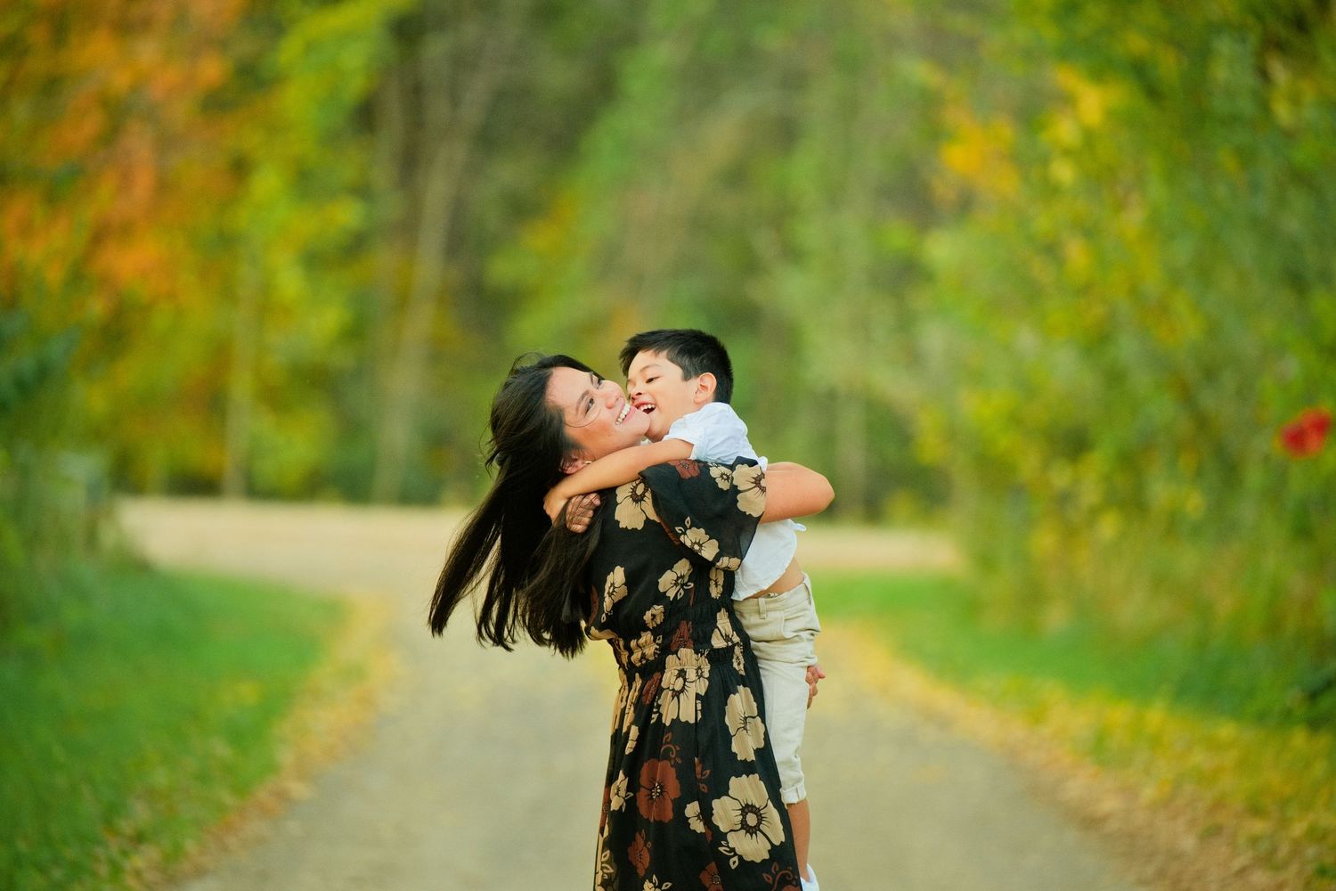 A joyful mother lifts her laughing toddler on a scenic tree-lined path during autumn.