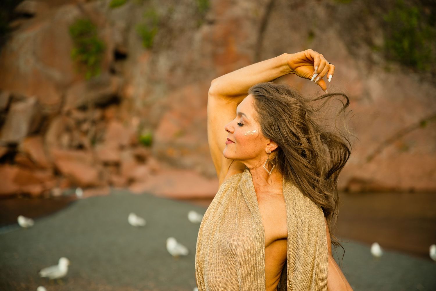 Woman with long brown hair and a raised arm poses in a golden shawl on a rocky beach with white seagulls nearby.