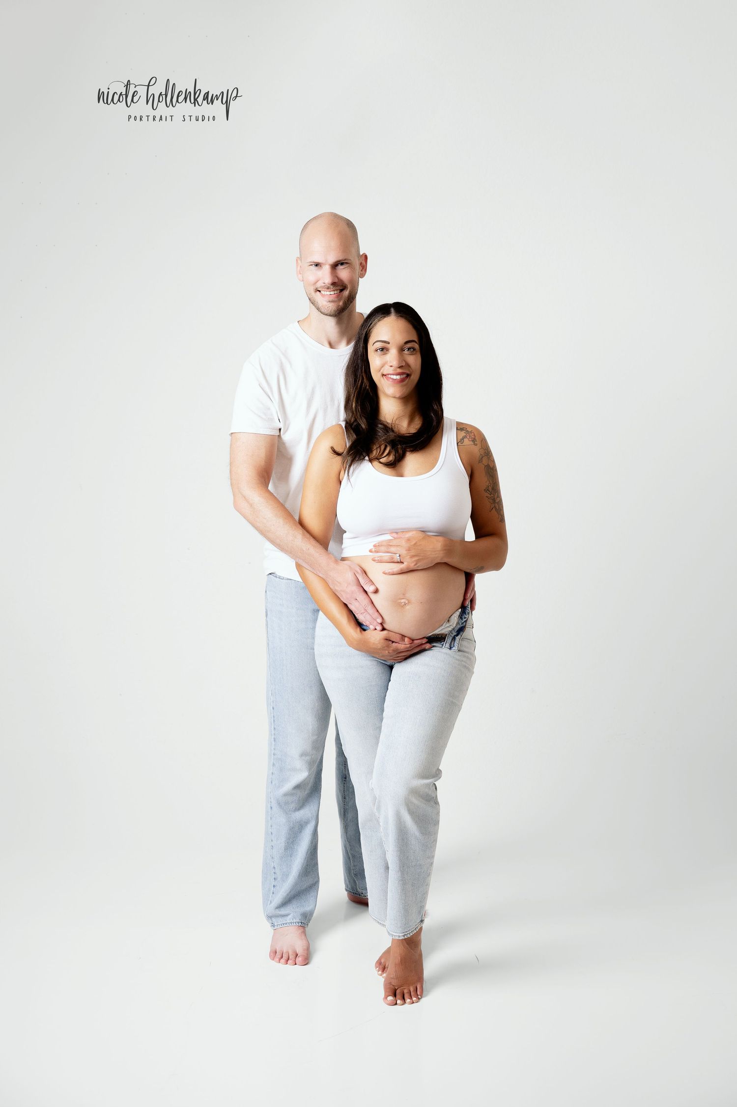 Couple poses for maternity portrait, woman in white crop top and jeans cradling baby bump, man in white shirt behind her.