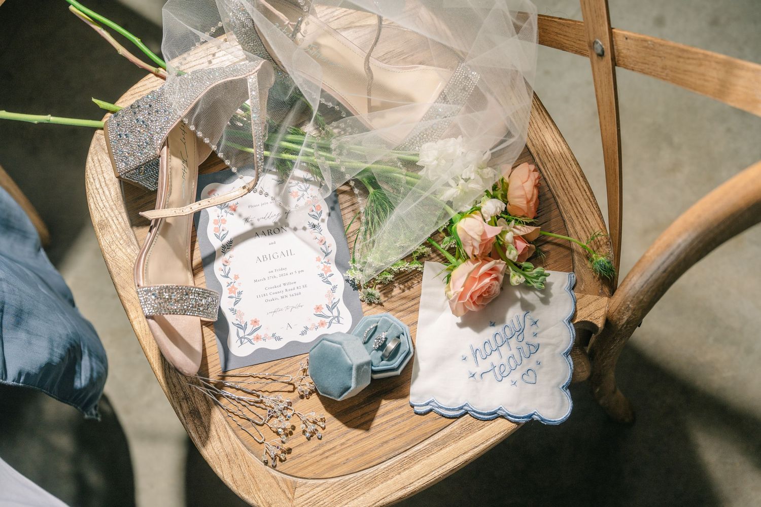 crooked willow weddings Wedding flat lay with invitation, veil, pink tulips, blue ring box, and handkerchief on wooden chair.