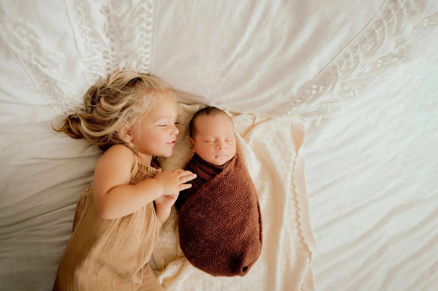 Two children lying together on white bedding during sweet portrait session.
