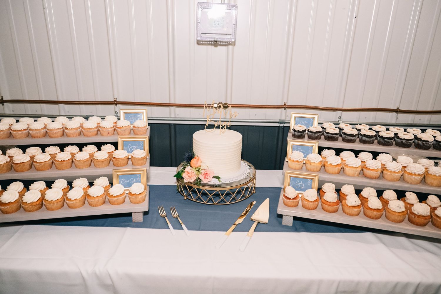 Elegant wedding dessert table with a two-tier white cake surrounded by rows of frosted cupcakes on wooden display stands.