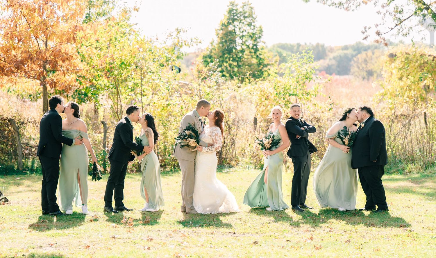 Wedding party kisses and embraces on a sunny autumn day with colorful fall foliage in the background.
