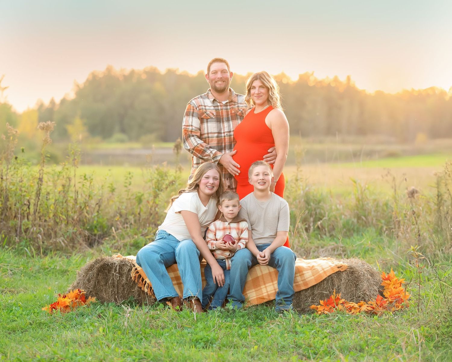 Family poses together outdoors in autumn field at sunset with pumpkins and fall decorations.