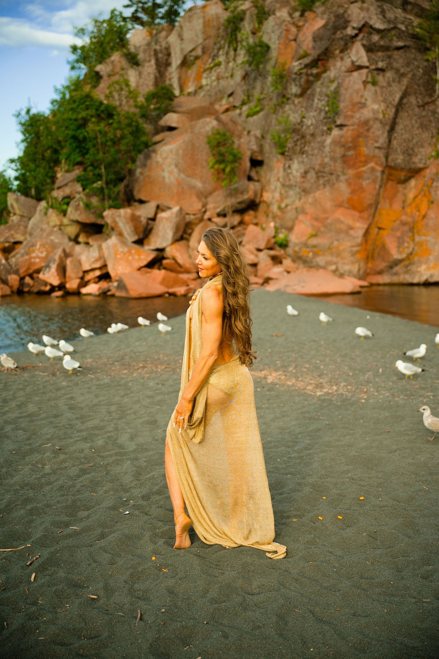 Woman in golden dress stands on dark sand beach with seagulls near red rock cliffs.
