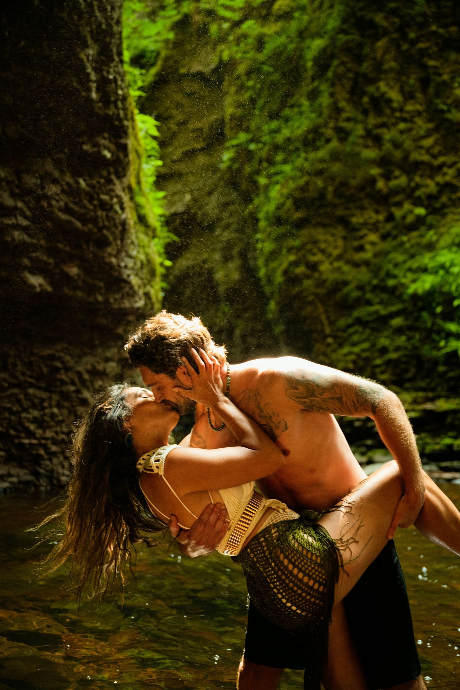 A passionate couple kissing in a lush green canyon waterfall, man lifting woman in a romantic embrace.