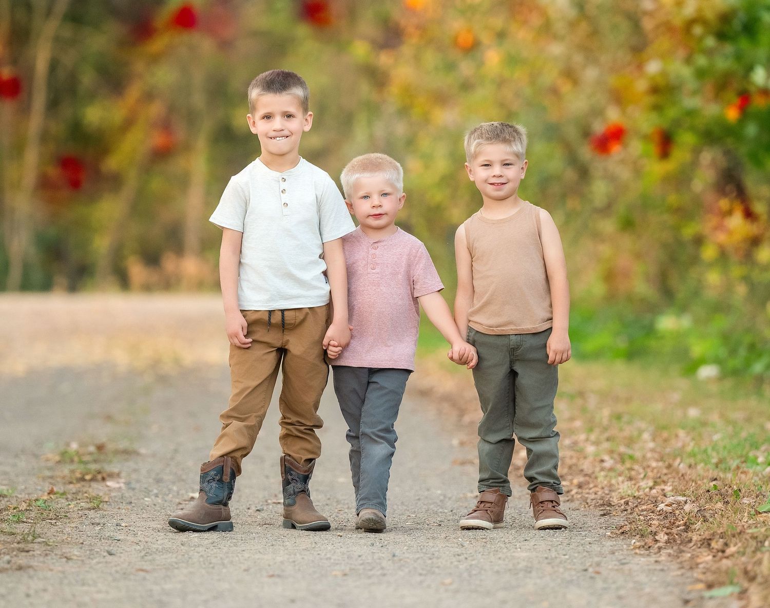 Three children walk down an autumn path surrounded by colorful fall foliage and red flowers.