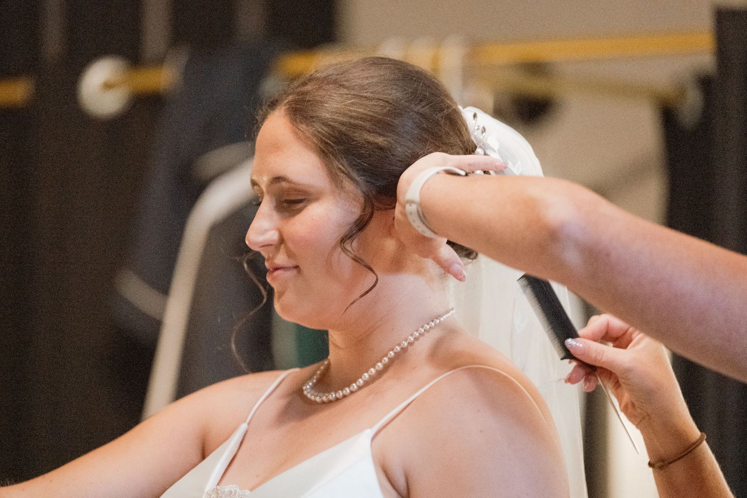 A bride gets help putting on her pearl necklace while getting ready for her wedding ceremony.